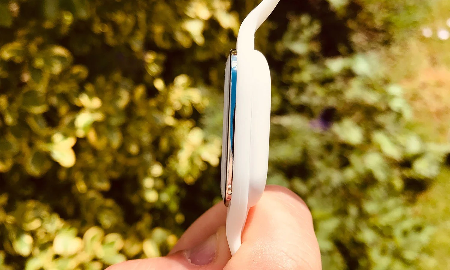 The image shows a side view of a Moskitopro bracelet held in hand. The bracelet is thin and white, with a slightly curved design and a silver case visible on the side. The blurred background suggests an outdoor setting, with green leaves in the background. The focus is on the device's slimness and elegance, which appears light and discreet.