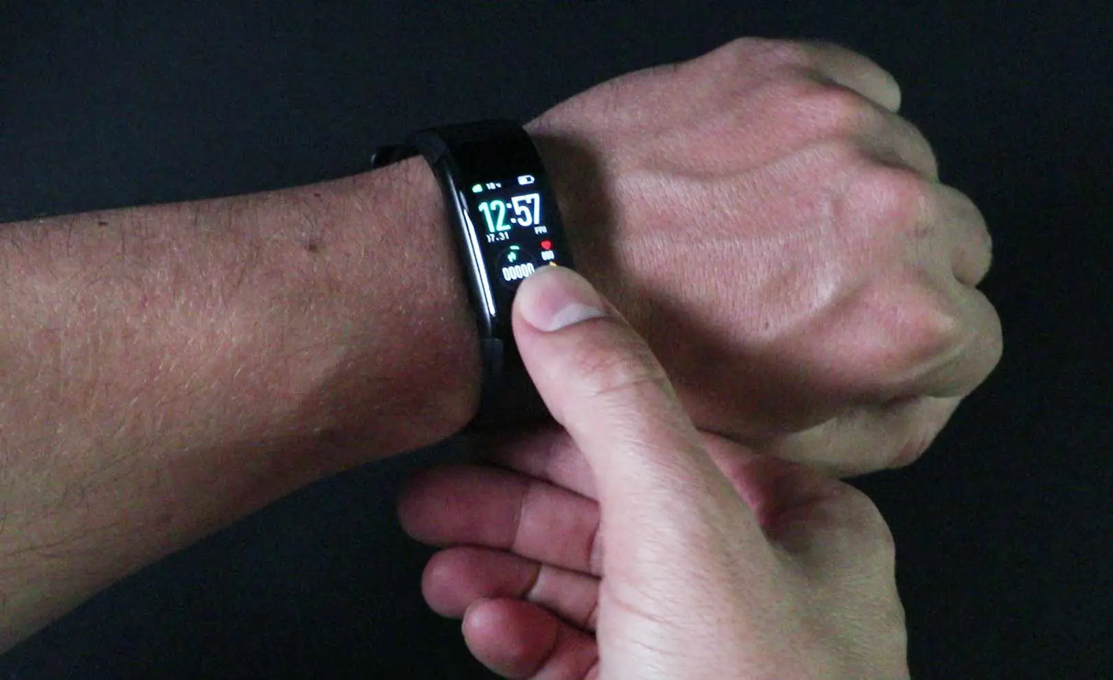 This image shows a hand wearing a black SmartTrack watch displaying the time '12:57' on a small screen. Another hand interacts with the watch's touchscreen by pressing on it. The watch also shows icons such as the step count, date, and battery status.